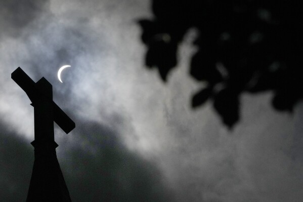 Clouds part as a partial eclipse of the sun and moon is seen atop the cross on the steeple of the New Sweden Evangelical Lutheran Church Monday, April 8, 2024, in Manor, Texas. (AP Photo/Charles Rex Arbogast)
