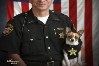 This 2006 image provided by John Hoffart shows then Sheriff Dan McClelland and his small police dog Midge at the Geauga County, Ohio, sheriff's department. Both died on Wednesday, April 14, 2021. McClelland after a lengthy battle with cancer and Midge, perhaps, of a broken heart. The family said they will be buried together. McClelland retired in 2016, after 13 years as sheriff, and 44 total in the department. The last ten with Midge, a drug-sniffing Chihuahua-rat terrier mix certified by Guinness World Records as the smallest police dog on the globe. (John Hoffart via AP)