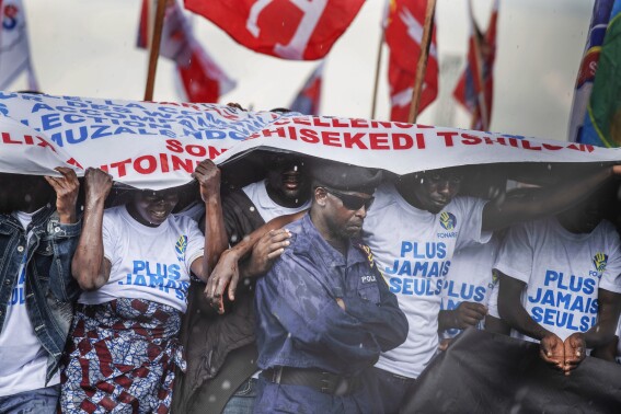 Supporters take cover from the rain under a banner as they wait for the arrival of President Felix Tshisekedi, who is seeking re-election, at a campaign rally in Goma, Congo, Sunday, Dec. 10, 2023. (AP Photo/Moses Sawasawa)