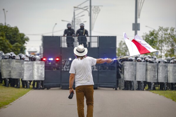 FILE - Artist Ales Pushkin, who died in July while imprisoned in Belarus after human rights activists say authorities deprived him of medical care, waves a red-and-white flag that symbolizes opposition to President Alexander Lukashanko in front of a police blockade during a protest in Minsk, Belarus, on Sunday, Aug. 23, 2020. Tens of thousands of demonstrators turned out following the widely disputed election that gave Lukashenko another term in office. (AP Photo/File)