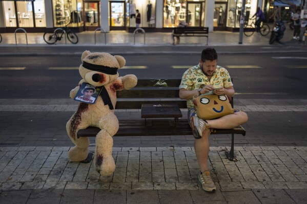 A man sits next to an installation of a blindfolded giant teddy bear in Tel Aviv, Israel, Monday, Nov. 6, 2023. The installation is meant to draw attention to over 240 people who were abducted during the Hamas cross-border attack on Oct. 7. (AP Photo/Bernat Armangue)