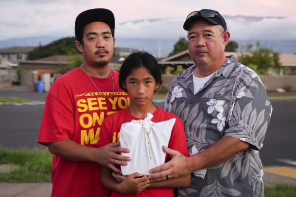 Briena Mae Rabang, 10, holds the ashes of her great-grandmother Sharlene Rabang, who was named as the 100th victim of the Lahaina wildfire, while posing for a photo with her father Branden, left, and grandfather Brandon, right, Friday, Dec. 8, 2023, in Kahului, Hawaii. Sharlene's family fought to have her listed as a victim due to smoke inhalation after she died weeks after fleeing the fire. "Me and my mom was really close, we talked multiple times a day," Brandon said. (AP Photo/Lindsey Wasson)