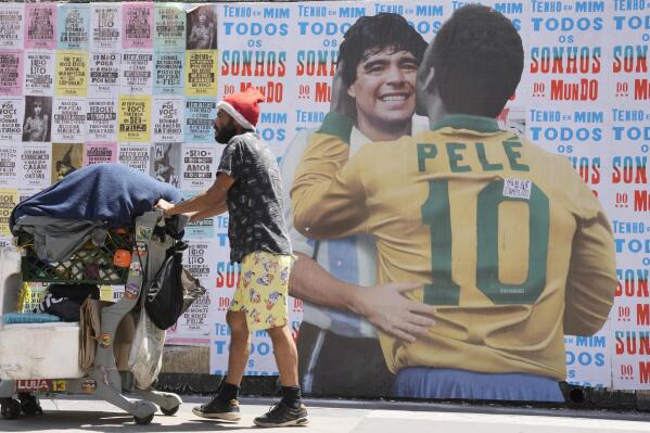 A man pushes a cart loaded with garbage for recycling in front of a mural depicting Brazilian soccer legend Pele embracing late Argentinean soccer star Diego Maradona in Sao Paulo, Brazil, Saturday, Dec. 24, 2022. Pele is hospitalized to continue his cancer treatment. (AP Photo/Andre Penner)