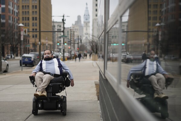 Temple University doctoral student Jaggar DeMarco poses for a portrait while utilizing a battery powered ventilator in Philadelphia, Wednesday, March 6, 2024. “I’m constantly angry that my life and what I can do with (it) is sometimes determined by insurance companies and bureaucracy,” says DeMarco, who has chronic respiratory failure. (AP Photo/Matt Rourke)