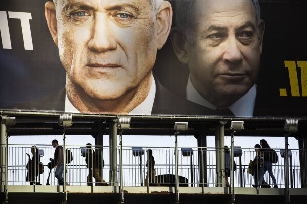 FILE - In this Feb. 18, 2020 file photo, People walk on a bridge under an election campaign billboard for the Blue and White party, the opposition party led by Benny Gantz, left, in Ramat Gan, Israel.  Prime Minister Benjamin Netanyahu of the Likud party is pictured at right. Israel finds itself in a familiar place after a tumultuous election campaign, with iconoclastic politician Avigdor Lieberman seemingly in control of the country’s fate.   Opinion polls ahead of the March 2 vote show that neither Netanyahu nor his challenger Gantz will be able to form a coalition government without him.  Lieberman remains cagey about his intentions, raising the possibility his brinkmanship could end up forcing yet another election.(AP Photo/Oded Balilty)