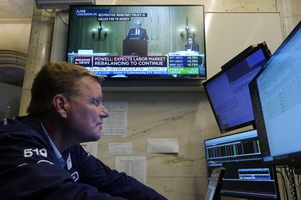 Trader John Bowers works on the floor of the New York Stock Exchange, Friday, Aug. 25, 2023, as Federal Reserve Chair Jerome Powell's speech shows on a television screen. Stocks are holding on to gains after Powell said more rate hikes could be on the way to continue the Fed's fight against inflation. (AP Photo/Richard Drew)