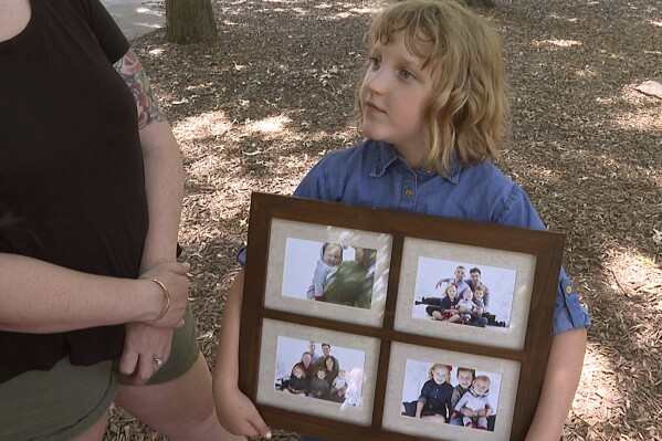 Olive Wilson shows off her family photo as she plays at a park on Friday, June 14, 2024. The now 7-year-old posed for the pictures, taken in 2021, with her two siblings, two mothers, the sperm donor who acts as a godfather figure and his husband. On Sunday, this family will celebrate Father's Day together. "He cares about us a lot," she says. (AP Photo/Nick Ingram)