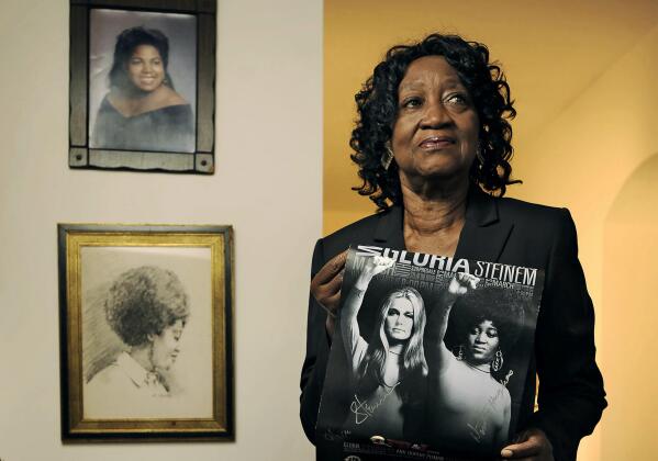 Dorothy Pitman Hughes poses in her St. Johns, Fla., home on Sept. 24, 2013, with a poster using a 1970's image of herself and Gloria Steinem. Hughes, a pioneering Black feminist, child welfare advocate and activist who formed a powerful speaking partnership with Steinem and appeared with her in one of the most iconic photos of the feminist movement, has died. Hughes died Dec. 1, 2022, in Tampa, Fla. She was 84. (Bob Self/The Florida Times-Union via AP)