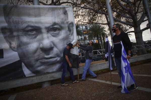 In this Saturday, April 25, 2020 photo, demonstrators wearing protective face masks amid concerns over the country's coronavirus outbreak, hang a banner showing Israeli Prime Minister Benjamin Netanyahu during "Black Flag" protest against Netanyahu and government corruption, at Rabin Square in Tel Aviv, Israel. During the past two weekends, thousands of people have gathered in perfect geometric patterns to comply with public-health restrictions as they express their anger over the continued rule of a prime minister charged with serious crimes. (AP Photo/Oded Balilty)