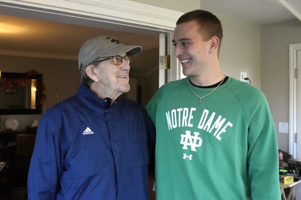 Saline High School quarterback CJ Carr, right, stands next to his grandfather Lloyd Carr, left, Nov. 14, 2023 in Saline, Mich. Carr will be among the hundreds of football players to sign a national letter of intent this week, sealing his commitment to join Notre Dame, but no one has a story quite the same. (AP Photo/Carlos Osorio)