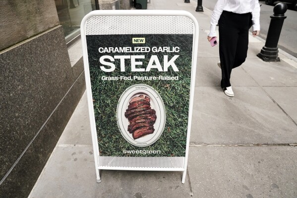A person walks past a sign for Sweetgreen's new caramelized garlic steak bowl on Thursday, May 9, 2024, in New York. The announcement of Sweetgreen that it’s adding beef to its menu led to strong reactions online, with customers questioning the company’s carbon neutral plans. (AP Photo)