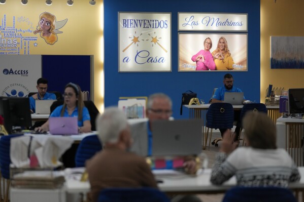 An image of the "Insurance Godmothers" Odalys Arevalo, right, and Mercy Cabrera, hangs on the wall as insurance agents work at Las Madrinas de los Seguros (Spanish for "The Godmothers of Insurance," a health insurance agency serving Spanish-speaking clients, at a shopping center in Miami, Tuesday, Dec. 5, 2023. (AP Photo/Rebecca Blackwell)