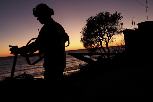 Los Angeles Fire Department's Mike Alvarez works on extinguishing hot spots in the aftermath of the Palisades Fire as a Malibu resident, top right, watches the sunset from atop his beachfront home along the Pacific Coast Highway in Malibu, Calif., Sunday, Jan. 12, 2025. (Scott Strazzante/San Francisco Chronicle via AP)
