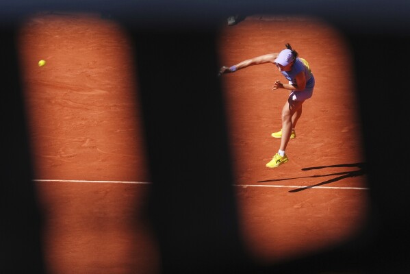 Poland's Iga Swiatek is seen through a fence as she serves against Romania's Jaqueline Cristian during their third round match of the French Tennis Open, at the Roland-Garros stadium, in Paris, Friday, May 30, 2025. (AP Photo/Lindsey Wasson)
