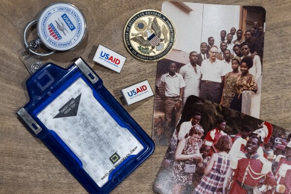 Anna Eisenberg's empty USAID badge holder is seen next to USAID pins, a coin from the USAID Office of Inspector General awarded to her mother, Cathy Votaw, and photographs of her grandfather, Albert Votaw, and his family during his service with USAID in West Africa circa 1968, Thursday, Feb. 27, 2025, in Washington. (AP Photo/Jacquelyn Martin)