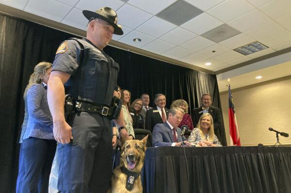 Georgia State Trooper Trent Kirchhefer stands with K-9 officer Vilma as Gov. Brian Kemp signs a bill clarifying penalties for harming a police dog, Wednesday, April 26, 2023, at Lake Lanier Islands in Buford, Ga. Kemp signed a number of public safety bills including one stiffening penalties for gang crimes and recruiting gang members. (AP Photo/Jeff Amy)