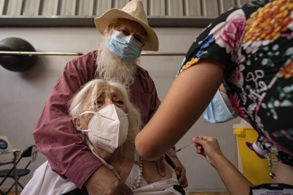 María Navarro, de 90 años, acompañada de su hijo Iván Moya, recibe una cuarta dosis de la vacuna de Pfizer contra COVID-19, su segundo refuerzo contra el nuevo coronavirus, en una instalación deportiva en Santiago de Chile, el lunes 7 de febrero de 2022. (AP Foto/Esteban Felix)
