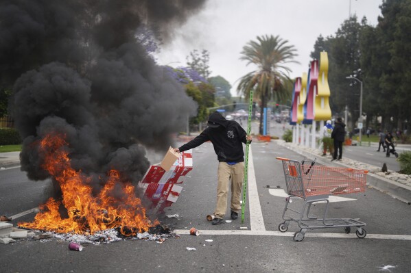 A protester places debris in a fire as Border Patrol personnel in riot gear and gas masks stand guard outside an industrial park in the Paramount section of Los Angeles, on Saturday, June 7, 2025 (AP Photo/Eric Thayer)