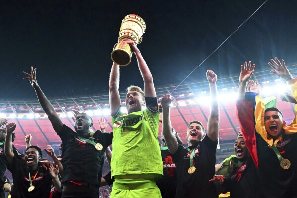 Leverkusen goalkeeper Lukas Hradecky holds up the trophy while his teammates celebrate winning the German Soccer Cup final match between 1. FC Kaiserslautern and Bayer Leverkusen at the Olympic Stadium in Berlin, Germany, Saturday, May 25, 2024. (Federico Gambarini/dpa via AP)