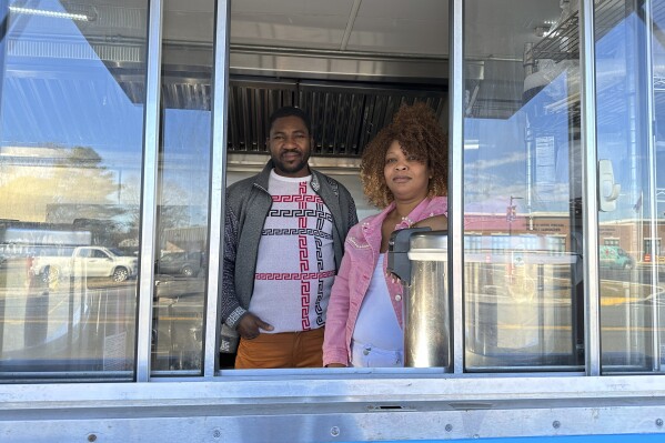 Theslet Benoir and Clemene Bastien stand at the window of their Eben-Ezer Haitian food truck in Parksley, Va., on Wednesday, Jan. 24, 2024. The married couple is suing the town in federal court over allegations that their food truck was forced to close. The couple also says a town councilman cut the mobile kitchen's water line and screamed, "Go back to your own country!" (AP Photo/Ben Finley)