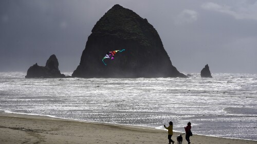 FILE - Beachgoers walk a dog and fly a kite as they near Haystack Rock, April 4, 2022, in Cannon Beach, Ore. Cannon Beach, a popular tourist destination, reopened Monday, July 17, 2023, after closing due to a cougar sighting near the iconic Haystack Rock. The U.S. Fish and Wildlife Service said Monday the cougar had moved on, as confirmed by wildlife and law enforcement officials. (AP Photo/Charles Rex Arbogast, File)