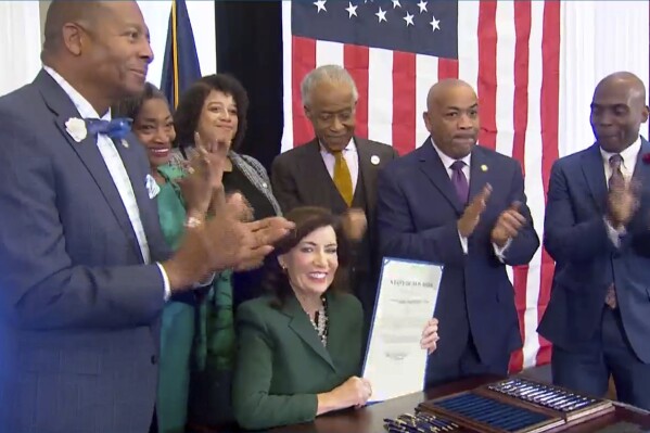 CORRECTS CITY TO NEW YORK - This image from video provided by the Office of The Governor shows New York Gov. Kathy Hochul, seated center, holds a bill she signed in New York, Tuesday, Dec. 19, 2023, to create a commission tasked with considering reparations to address the persistent, harmful effects of slavery in the state. She is joined by, standing from left: Dr. Yohuru Williams, Founding Director of the Racial Justice Initiative at the University of St. Thomas; Andrea Stewart-Cousins, Majority Leader of the NY State Senate; Michaelle Solages, NY State Assembly Woman; Rev. Al Sharpton; Carl Heastie, Speaker of the NY State Assembly; James Sanders, NY State Senator. (Office of the Governor via AP)