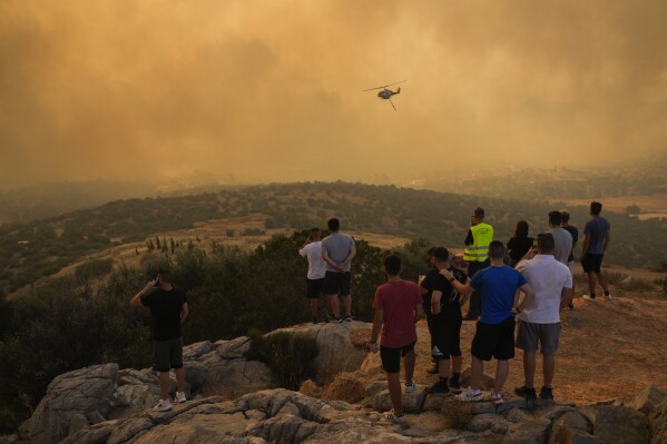 A firefighting helicopter flies through smoke as people look on in Mandra west of Athens, on Tuesday, July 18, 2023. In Greece, where a second heatwave is expected to hit Thursday, three large wildfires burned outside Athens for a second day. Thousands of people evacuated from coastal areas south of the capital returned to their homes Tuesday when a fire finally receded after they spent the night on beaches, hotels and public facilities. (AP Photo/Petros Giannakouris)