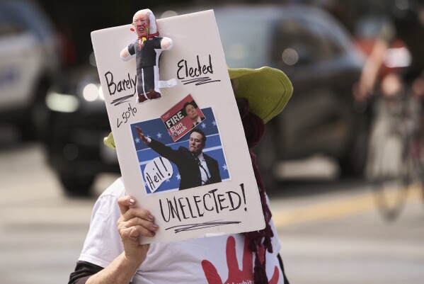 A protestor hold a banner during a protest outside a Tesla showroom against U.S. President Donald Trump and Tesla CEO Elon Musk outside a Tesla showroom in Santa Monica, Calif., on Saturday, March 29, 2025. (AP Photo/Damian Dovarganes)