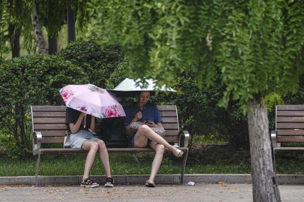 Residents carry umbrellas to shield from the sun as they take rest on a bench on a hot day in Beijing, Monday, July 3, 2023. Heavy flooding has displaced thousands of people around China as the capital had a brief respite from sweltering heat. (AP Photo/Andy Wong)