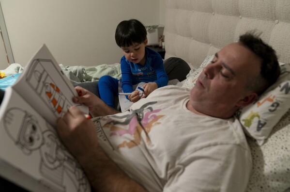 Scott Quinn and his 4-year-old son, Ethan, work on coloring books in a bed in Concord, Calif., Wednesday, Nov. 1, 2023. Ethan's parents are among those who have opted for a private daycare instead of a free “transitional kindergarten.” Quinn said he has been discouraged to see Ethan — one of the oldest kids in his daycare class — pick up the behavior of kids who are several years younger than him. “In retrospect, it would have been better to send him to school to be around kids his age and older,” he said. (AP Photo/Jae C. Hong)