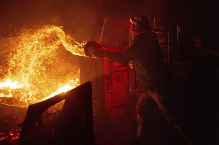 Francis Lopez douses flames with a bucket of water as a wildfire called the Highland Fire burns through his property in Aguanga, Calif., Monday, Oct. 30, 2023. A wildfire fueled by gusty Santa Ana winds ripped through rural land southeast of Los Angeles on Monday, forcing thousands of people from their homes, fire authorities said. (AP Photo/Ethan Swope)