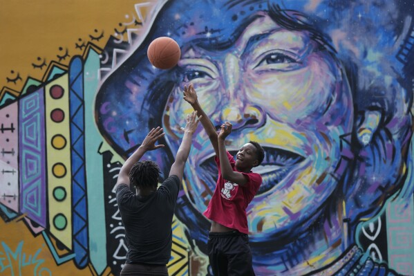 Jamari Baugh, 15, shoots a basketball past a friend as they play on a court next to a mural of author Zora Neale Hurston, who grew up in Eatonville and depicted a fictionalized version of the historic Black town in her work, Wednesday, Aug. 23, 2023, at Elizabeth Park in Eatonville, Fla. Descendants of the community work to boost its economy and preserve the local heritage and culture, put on display at the town's annual ZORA! Festival (AP Photo/Rebecca Blackwell)