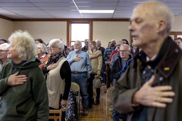 Residents stand for the Pledge of Allegiance at the start of the annual Town Meeting in Elmore, Vt., Tuesday, March 5, 2024. Town Meeting is a tradition that, in Vermont, dates back more than 250 years, to before the founding of the republic. But it is under threat. Many people feel they no longer have the time or ability to attend such meetings. Last year, residents of neighboring Morristown voted to switch to a secret ballot system, ending their town meeting tradition. (AP Photo/David Goldman)