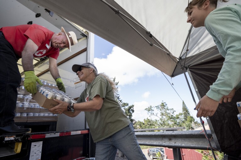 Volunteers of American Red Cross, Tom Hobbs, of Colorado, left, hands a box of cans of water to Kimberly Patrick-Chapman, of Michigan, at the Kula distribution hub on Wednesday, Sept. 27, 2023, in Kula, Hawaii. (AP Photo/Mengshin Lin)