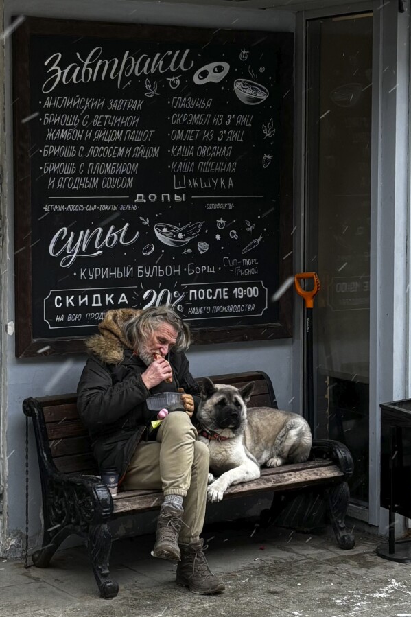 A man eats a sandwich while sitting on a bench with his dog in Moscow, Russia, Thursday, Feb. 13, 2025. (AP Photo/Pavel Bednyakov)