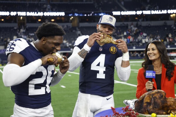 Dallas Cowboys cornerback DaRon Bland, left, and quarterback Dak Prescott (4) celebrate by eating Thanksgiving turkey legs next to reporter Tracy Wolfson after the Cowboys defeated the Washington Commanders in an NFL football game Thursday, Nov. 23, 2023, in Arlington, Texas. (AP Photo/Michael Ainsworth)