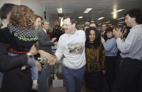 FILE - Wearing a "Hello World" sweatshirt printed with his picture, former hostage Terry Anderson greets happy colleagues, Dec. 10, 1991, at The Associated Press headquarters in New York, as he walks with his arm around fiancée Madeleine Bassil, center right. Anderson, the globe-trotting Associated Press correspondent who became one of America’s longest-held hostages after he was snatched from a street in war-torn Lebanon in 1985 and held for nearly seven years, died Sunday, April 21, 2024. He was 76. (AP Photo/Ron Frehm, File)