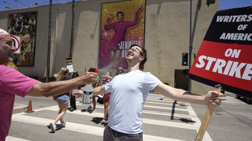 Luke DePalatis, right, gets a cooling spritz of water from Michael Abel during a rally by striking writers and actors outside Warner Bros. studios Friday, July 14, 2023, in Burbank, Calif. Both are with the WGA. This marks the first day actors formally joined the picket lines, more than two months after screenwriters began striking in their bid to get better pay and working conditions and have clear guidelines around the use of AI in film and television productions. (AP Photo/Mark J. Terrill)