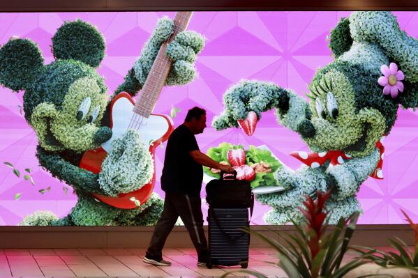 A traveler walks past a jumbo screen projecting images of Walt Disney World in the east hall atrium at Orlando International Airport, Thursday, May 25, 2023 in Orlando, Fla. Auto club AAA said this summer could be “one for the record books, especially at airports,” with more than 43 million Americans projected to travel 50 miles or more. (Joe Burbank /Orlando Sentinel via AP)