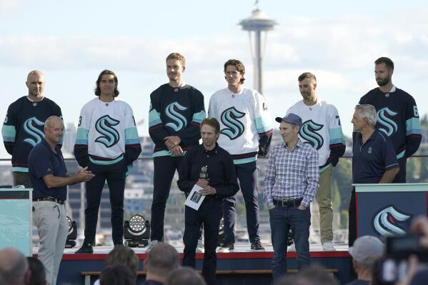 New Seattle Kraken NHL hockey players, back row from left, Mark Giordano, Brandon Tanev, Jamie Oleksiak, Hadyn Fluery, Jordan Eberle and Chris Dreidger stand on stage with Kraken owners David Wright, front left, Jerry Bruckheimer, front center, and Andy Jassy, front second from right, and Kraken general manager Ron Francis, front right, Wednesday, July 21, 2021, after being introduced during the Kraken's expansion draft event in Seattle. Jassy is also president and CEO of Amazon.com. (AP Photo/Ted S. Warren)