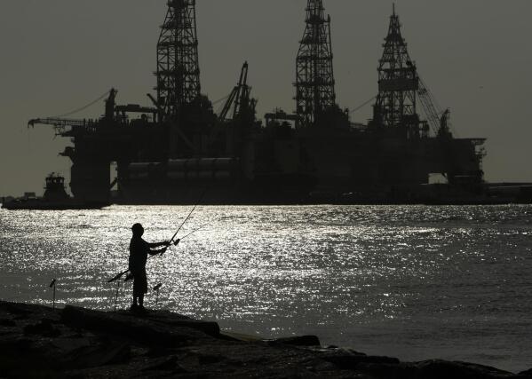 FILE - A man wears a face mark as he fishes near docked oil drilling platforms, Friday, May 8, 2020, in Port Aransas, Texas. The U.S. Interior Department on Wednesday, Nov. 17, 2021, is auctioning vast oil reserves in the Gulf of Mexico estimated to hold up to 1.1 billion barrels of crude. It's the first such sale under President Joe Biden and underscores the challenges he faces to reach climate goals that rely on cuts in fossil fuel emissions.  (AP Photo/Eric Gay, File)