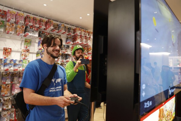 Darren Stallcup, left, of San Francisco reacts as Nicholas Oehlberg, right, of San Francisco documents the event as they play the Mario Kart World for Nintendo Switch 2 game with others at the Nintendo Store for a pre-launch event for the Ninentdo Switch 2 on Wednesday, June 4, 2025, in San Francisco. (Lea Suzuki/San Francisco Chronicle via AP)