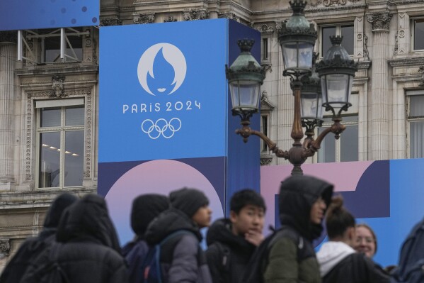 Youth gather at Paris City Hall where a Paris 2024 Olympic sign is displayed in Paris, Tuesday, March 5, 2024. The French government announced that tourists won't be allowed to watch the opening ceremony of the Paris Olympics for free as initially promised, the government announced Tuesday, as it grapples with security concerns about the unprecedented open-air event along the Seine River. (AP Photo/Michel Euler)