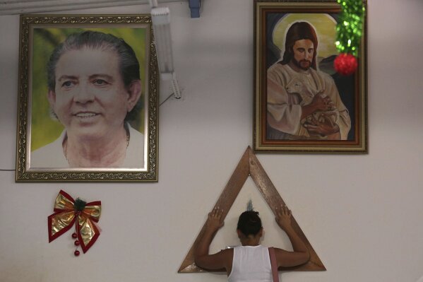 
              In this Jan. 4, 2019 photo, a woman places her hands in prayer on a wooden triangle, between framed images of spiritual healer Joao Teixeira de Faria and Jesus Christ, inside the Casa de Dom Inacio, in Abadiania, Brazil. Hundreds of women have accused de Faria of sexual abuse. The mounting accusations are turning the 77-year-old spiritual guru into Brazil's first major figure to go down in the #metoo era. (AP Photo/Eraldo Peres)
            