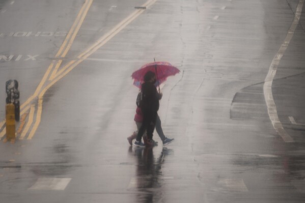 People cross the street with umbrellas in heavy rain in Los Angeles, Monday, Feb. 19, 2024. (AP Photo/Damian Dovarganes)