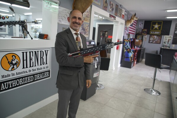 Army veteran and mayoral candidate Freddie de la Cruz poses with a semiautomatic shotgun he's raffling off at Fort Liberty Pawn & Gun, Friday, May, 9, 2025. (AP Photo/Allen G. Breed)