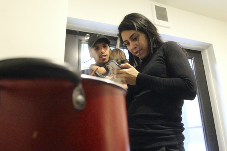 Barbara Peraza-Garcia looks astatine a telephone while cooking successful a communal room while her partner, Franklin Peraza holds their daughter, Frailys, successful a 'micro apartment' building successful Seattle connected Monday, March 11, 2024. (AP Photo/Manuel Valdes)