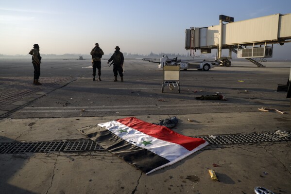 A Syrian flag lies on the ground as opposition fighters stand on the tarmac of the Aleppo international airport, Monday, Dec. 2, 2024.(AP Photo/Omar Albam)