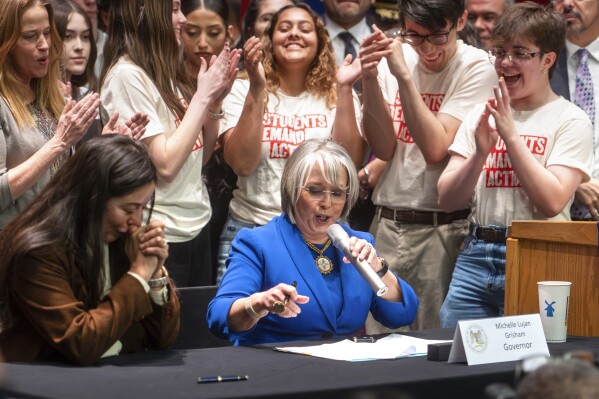Gov. Michelle Lujan Grisham, center, prepares to sign House Bill 129, as bill co-sponsor Rep. Andrea Romero, D-Santa Fe, left, clasps her hands at West Mesa High School in Albuquerque, New Mexico, Monday, March 4, 2024. Some guns will be banned at voting locations and the waiting period for purchasing firearms is being extended to seven days under measures signed into law Monday by Grisham. (Jon Austria/The Albuquerque Journal via AP)