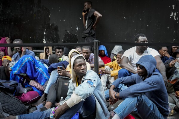 Migrants sit in a queue outside of The Roosevelt Hotel that is being used by the city as temporary housing, Monday, July 31, 2023, in New York. (AP Photo/John Minchillo)